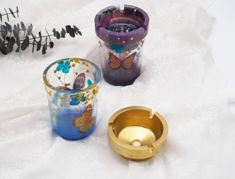 Puede incluir: Tres ceniceros decorativos. Dos son de resina transparente con dise&ntilde;os de mariposas y flores, uno con un fondo p&uacute;rpura y estrellado. El tercero es un cenicero redondo de metal dorado con un agujero central y tres muescas.