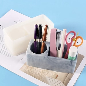 May include: A grey and white marbled desk organiser with two compartments. One holds pens, the other a stapler, scissors, and glue stick. A second, empty white organiser sits behind it. The items are on white paper.