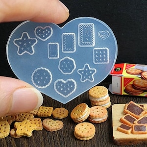 May include: A clear, heart-shaped silicone mold with various biscuit shapes, including stars, hearts, and rectangles. Miniature biscuits and sandwich cookies are arranged nearby, with a box of cookies in the background. The image showcases a baking or crafting theme.