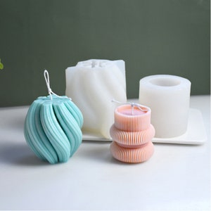 May include: A collection of decorative candles in various shapes and colors. One candle is a light teal spiral design, another is a light pink stacked design, and two white molds are in the background. The candles are on a white tray.
