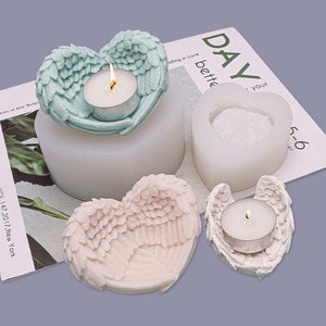 Pode incluir: Uma coleção de castiçais e moldes em forma de coração. Um castiçal verde-azulado com design de asas de anjo contém uma vela de chá acesa. Um molde branco e um castiçal rosa também são visíveis. A palavra "DAY" é visível em uma revista.