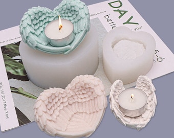 Puede incluir: Una colección de candelabros y moldes en forma de corazón. Un candelabro verde azulado con diseño de alas de ángel sostiene una vela de té encendida. También son visibles un molde blanco y un candelabro rosa. La palabra "DAY" es visible en una revista.