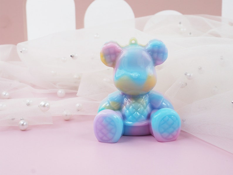 K&ouml;nnte beinhalten: Eine farbenfrohe B&auml;renfigur in Blau-, Lila-, Gelb- und Rosat&ouml;nen. Der B&auml;r hat ein Steppmuster auf Brust und F&uuml;&szlig;en. Er hat eine kleine Schlaufe zum Aufh&auml;ngen. Der Hintergrund ist ein zartes Rosa mit wei&szlig;em Stoff und Perlen.