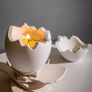 May include: A white, eggshell-shaped candle holder with a lit candle inside, and a matching empty eggshell-shaped bowl. The candle holder sits on a base of broken pieces. The text "Daisy Note Craft" is visible.