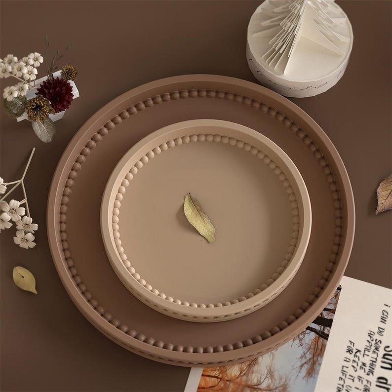 May include: A nested set of decorative trays in shades of brown and beige. The largest tray is a darker brown, with a beaded edge. The middle tray is a lighter beige, also with a beaded edge. A small, dried leaf rests on the middle tray.
