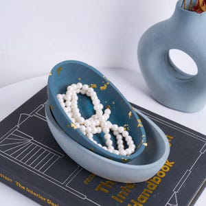 Op de afbeelding: Twee ovale decoratieve kommen, één teal en één lichtblauw, op elkaar gestapeld. De teal kom bevat een witte parelketting en goudblad accenten. Een blauwe vaas staat op de achtergrond.