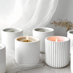 Peut inclure: Plusieurs bougies cylindriques blanches avec des mèches allumées. L'une des bougies présente un design côtelé. Les bougies sont posées sur une surface blanche avec un tissu blanc et doux en arrière-plan. Les flammes sont vives.