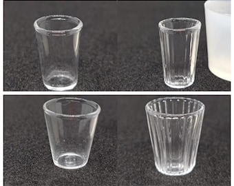May include: Four clear glass shot glasses. Two are cylindrical, and two are tapered. One cylindrical glass has vertical ridges. The glasses are empty and sit on a dark surface.