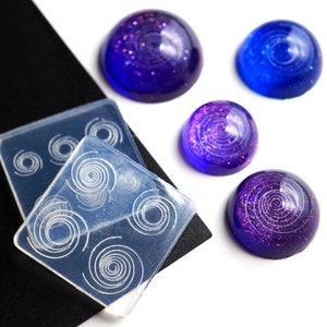 May include: A collection of resin art supplies, including a clear silicone mold with spiral designs and several finished resin pieces in shades of blue and purple. The resin pieces have a domed shape and glitter accents.