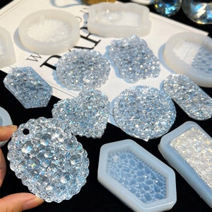 Può includere: Un assortimento di pezzi in resina trasparente e scintillante di varie forme, tra cui cerchi, cuori e rettangoli. Alcuni pezzi sono pieni, altri sono inseriti in stampi in silicone bianco. I pezzi hanno un aspetto glitterato e cristallino.