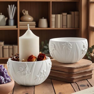 Pode incluir: Duas tigelas de cerâmica branca com desenhos florais em relevo. Uma tigela contém uma vela branca grande e flores secas. A outra está vazia. Ambas as tigelas estão sobre uma superfície de madeira, para decoração ou casa.