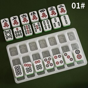 May include: A set of white and green Mahjong tiles with red and black markings, arranged on a clear plastic tray. The tiles feature Chinese characters and circular patterns. The background is a solid green color.