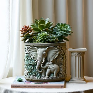 May include: A large, gray planter pot with an elephant relief design, filled with various green and reddish succulents. A small, decorative column stands beside the pot. The pot sits on a table with a book.