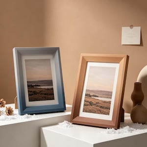 Puede incluir: Dos fotos de paisajes enmarcadas. Un marco es azul, el otro marrón. Ambos marcos son rectangulares con un soporte en la parte posterior. Las fotos representan un paisaje con un cuerpo de agua y campos.