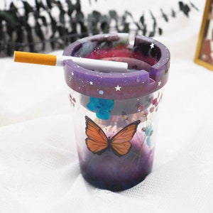 Puede incluir: Un cenicero de resina transparente con un borde morado salpicado de estrellas blancas. El cenicero presenta un dise&ntilde;o de mariposa y flores. Un cigarrillo encendido descansa en la ranura del cenicero.