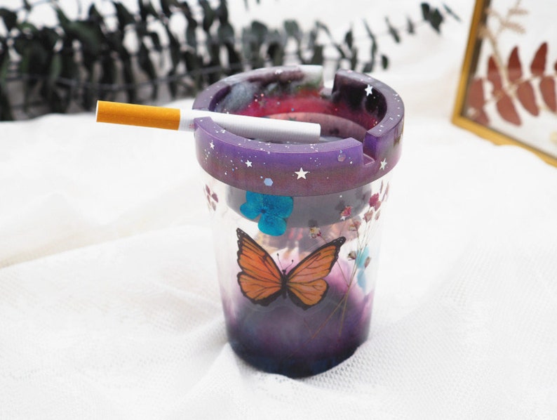 Puede incluir: Un cenicero de resina transparente con un borde morado salpicado de estrellas blancas. El cenicero presenta un dise&ntilde;o de mariposa y flores. Un cigarrillo encendido descansa en la ranura del cenicero.
