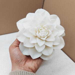May include: A large, white, artificial flower with multiple layers of petals. The flower is held in a person's hand, showcasing its intricate design and delicate texture. The petals are tightly arranged, creating a full, blooming appearance. The flower is a decorative item.