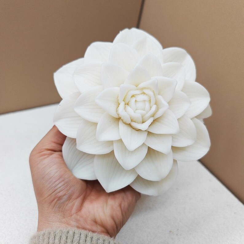 May include: A large, white, artificial flower with multiple layers of petals. The flower is held in a person's hand, showcasing its intricate design and delicate texture. The petals are tightly arranged, creating a full, blooming appearance. The flower is a decorative item.