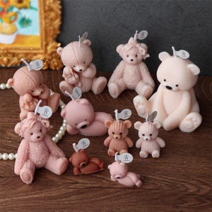 May include: A collection of teddy bear-shaped candles in various shades of pink and brown. The candles are in different poses, some sitting, some lying down, and some holding smaller bears. Each candle has a small tag attached.