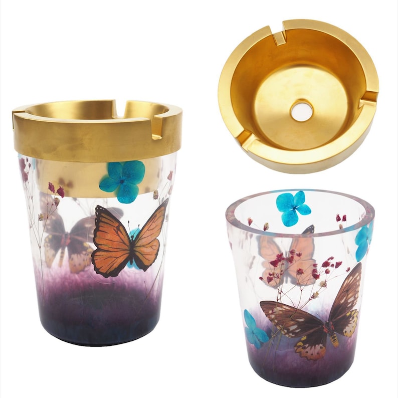 Puede incluir: Un juego de tres ceniceros decorativos. Dos son cil&iacute;ndricos con un borde dorado y dise&ntilde;os de mariposas y flores. El tercero es un cenicero circular de color dorado. Los ceniceros presentan mariposas, flores azules y detalles morados.