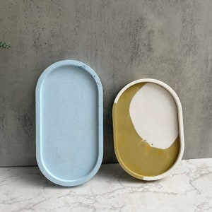 May include: Two oval-shaped concrete trays. One is light blue, the other is white and gold. The trays have rounded edges and are designed for decorative or functional use. The background is a neutral gray.