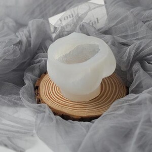 May include: A white, abstract-shaped silicone mold for crafting, resting on a small, round wooden slice. The mold is set against a backdrop of draped, sheer gray fabric, creating a soft, artistic presentation.