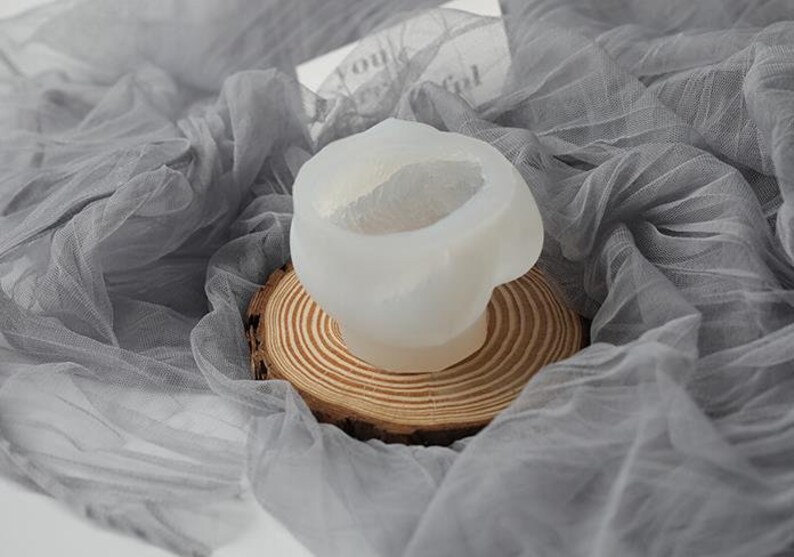 May include: A white, abstract-shaped silicone mold for crafting, resting on a small, round wooden slice. The mold is set against a backdrop of draped, sheer gray fabric, creating a soft, artistic presentation.