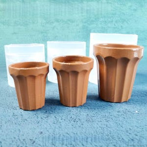 May include: Three terracotta-colored tea cups of varying sizes. The cups have a ridged design and are set against a blue background. Behind the cups are three translucent, rectangular molds.