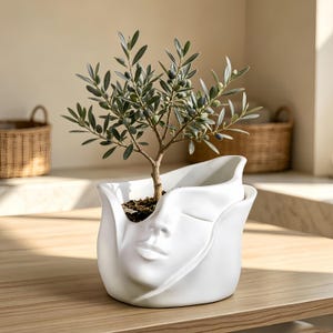 Op de afbeelding: Een witte keramische plantenbak in de vorm van een menselijk gezicht met gesloten ogen en volle lippen. De plantenbak bevat een kleine olijfboom met groene bladeren en kleine donkere olijven. Het oppervlak is glad en mat met gebogen randen.