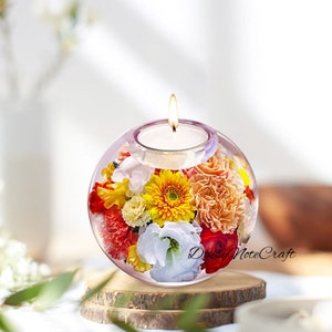 May include: A lit candle sits atop a clear, spherical glass candle holder filled with colorful flowers. The flowers include yellow, orange, red, and white blooms. The candle holder rests on a wooden base, with the text "Daisy NoteCraft" visible.