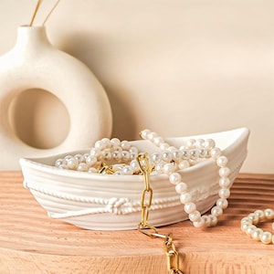 May include: A white ceramic boat-shaped jewelry dish filled with pearl necklaces and gold chain. The dish has a rope design around the rim. A white vase is in the background.