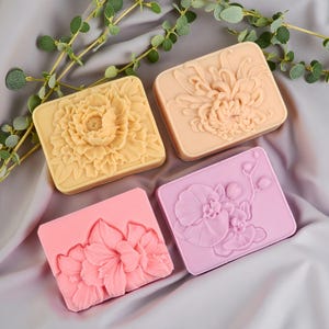 Pode incluir: Quatro moldes retangulares de silicone para sabonete ou cera, cada um com um design floral em relevo distinto. O conjunto inclui um molde de peônia amarela, um de crisântemo pêssego, um de lírio rosa e um de orquídea lilás.
