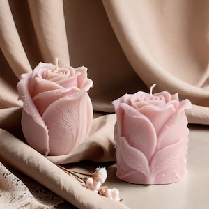 Puede incluir: Dos velas rosas con forma de rosa. Las velas son de color rosa suave y tienen diseños detallados de pétalos. Cada vela tiene una mecha blanca. Las velas están sobre una superficie de color neutro con un fondo de tela drapeada.