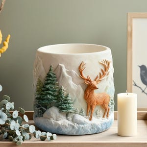 May include: A cream-colored decorative vase with a winter mountain scene and a brown deer. The vase features a relief design of snow-covered mountains, evergreen trees, and a standing deer. A white candle is next to the vase.