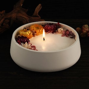 May include: A lit white candle in a low, white bowl, decorated with dried flowers in shades of yellow, red, and white. The candle's flame is visible. The background is dark, creating a contrast with the candle.