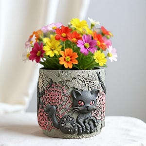 Può includere: Un vaso di fiori a tema Halloween con un design di gatto nero, ragnatele e pipistrelli. Il vaso è pieno di fiori colorati nei toni del rosso, arancione, giallo, rosa e bianco. Il vaso è grigio con dettagli rosa.