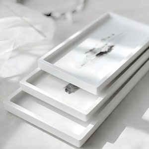 Könnte beinhalten: Ein Stapel von drei rechteckigen weißen Tabletts. Jedes Tablett hat einen erhöhten Rand und eine glatte Oberfläche. Das obere Tablett zeigt ein abstraktes Design in Schwarz und Grau. Die Tabletts sind übereinander gestapelt.