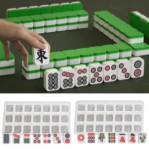 May include: A complete Mahjong set with green and white tiles, arranged on a green surface. The set includes tiles with various symbols and numbers, and two trays for storage. One tile is being held up, displaying a black character.