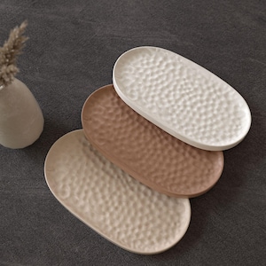 Könnte beinhalten: Drei ovale Servierplatten aus Keramik in Creme, Braun und Weiß. Jede Platte hat eine strukturierte, genoppte Oberfläche. Die Platten sind auf einer dunkelgrauen Oberfläche angeordnet, mit einer kleinen, cremefarbenen Vase und getrockneten Pflanzenstängeln im Hintergrund.