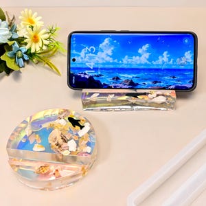 Puede incluir: Dos soportes para teléfono de resina. Uno es rectangular y sostiene un teléfono que muestra un paisaje marino. El otro es redondo, con conchas marinas y un pez negro incrustados.