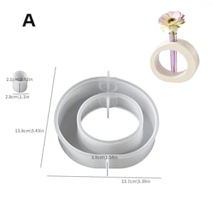 May include: A white, circular vase with a purple glass tube holding a single flower. Next to it is a gray, circular mold with a central ring. The mold measures 5.43 inches in diameter. A small, white, oval-shaped object is also shown.