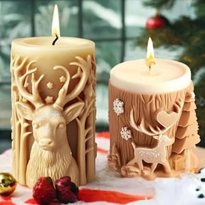 May include: Two lit, decorative candles. One is beige with a detailed deer and forest design. The other is brown with a deer and snowflake design. Both candles are on a white surface with Christmas decorations in the background.