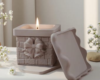 May include: A lit, square candle in a light gray container with a brick pattern and a bow design. The candle has a matching lid. The candle is on a white surface, with white flowers in the background.