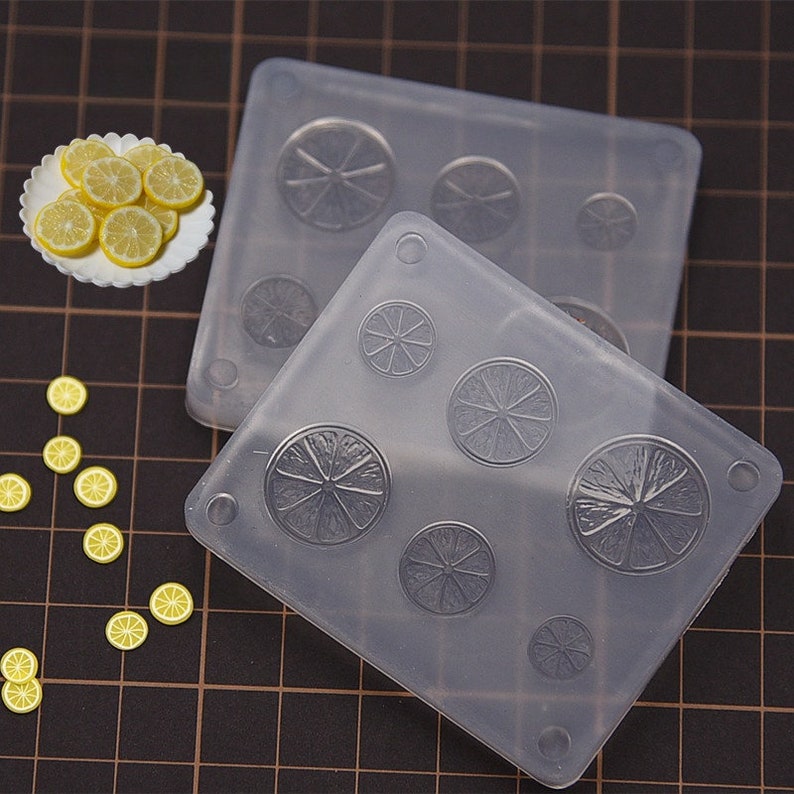 May include: Two clear silicone molds with lemon slice designs, alongside a plate of lemon slices. The molds have various sized circular impressions. Scattered around are small, yellow lemon slice embellishments. The background is a grid.
