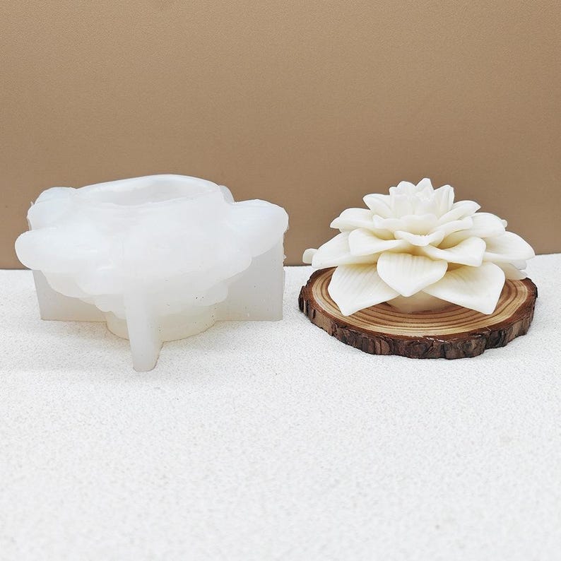 May include: A white silicone mold shaped like a flower and a finished white flower candle on a wooden slice. The candle has multiple layers of petals. The wooden slice has a natural, brown, circular shape.