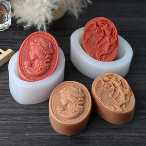 Puede incluir: Jabones ovalados en tonos coral y beige, con diseños de camafeos detallados de un perfil de mujer y un ángel. Los jabones se exhiben en moldes de silicona blancos, mostrando los intrincados patrones en relieve. Un artículo artesanal.