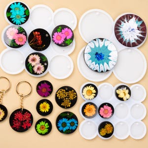 以下が含まれることがあります： さまざまな色のドライフラワーを使用した、手作りの樹脂製キーホルダーとコースターのコレクション。キーホルダーはゴールドカラーの金具を使用。コースターとキーホルダーは丸型で、白い型に入っています。