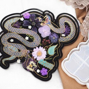 May include: A black resin coaster with a snake design, filled with dried flowers in shades of purple, white, and teal. The snake is blue with gold accents. A white silicone mold is in the background.