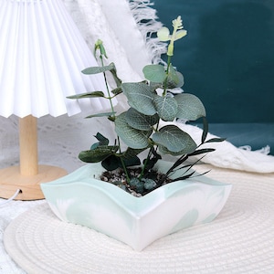 May include: A square, light green and white marbled ceramic planter holding a faux eucalyptus plant. The plant has green leaves and dark green stems. The planter is sitting on a white surface.
