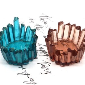 May include: Two crystal-shaped resin bowls. One is teal with glitter, the other is a peach color. Both have a crystal-like design around the rim. The bowls are on a white surface with cursive writing.
